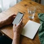 Pessoa com o celular na mão ao lado de um caderno, sobre a mesa de trabalho